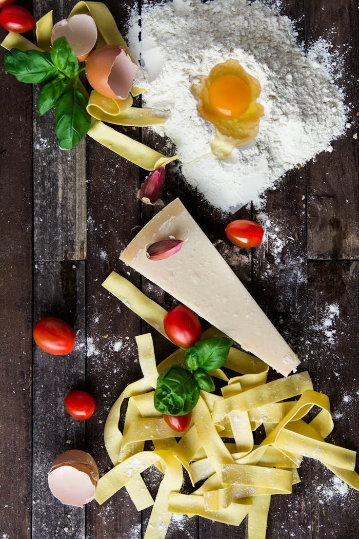 Top view of fresh pasta ingredients including flour, egg, parmesan, cherry tomatoes, and basil on a rustic wooden surface.
